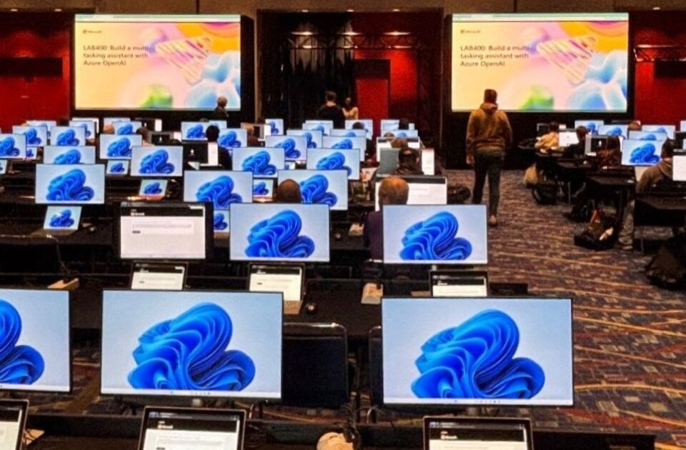 Ein Konferenzraum mit sehr vielen Arbeitsplätzen; einige Menschen sitzen an den Arbeitsplätzen und schauen auf die Monitore